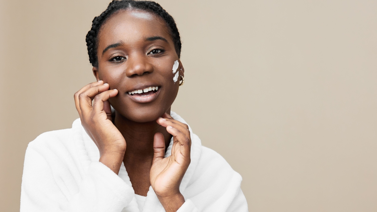 Smiling Black woman with natural skincare, wearing a white robe and facial cream, against a soft